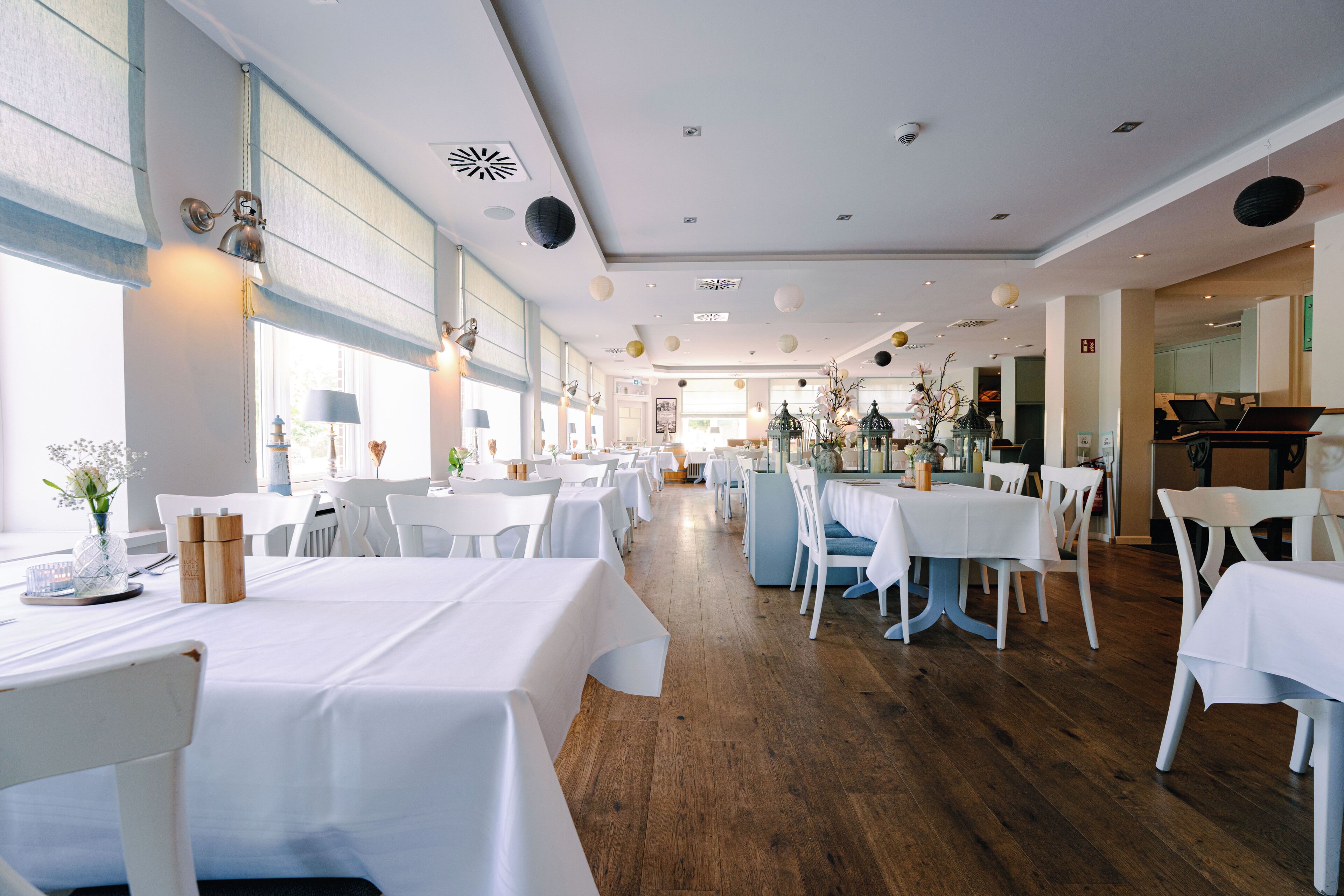 A spacious restaurant with white tables and chairs, large windows, and a wooden floor.