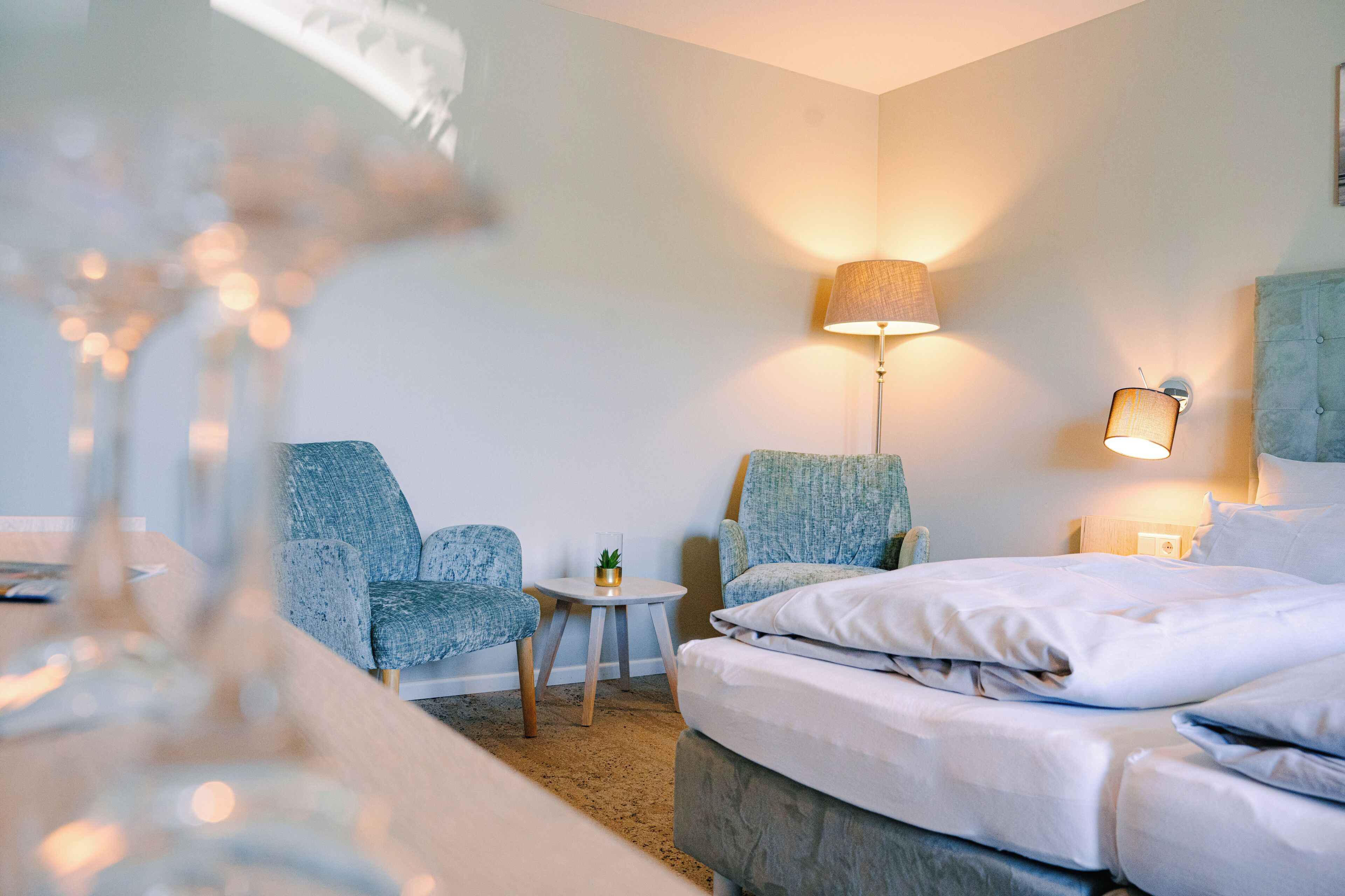 A hotel room with a bed, two armchairs, a small table with a lamp, and a wall-mounted light fixture.