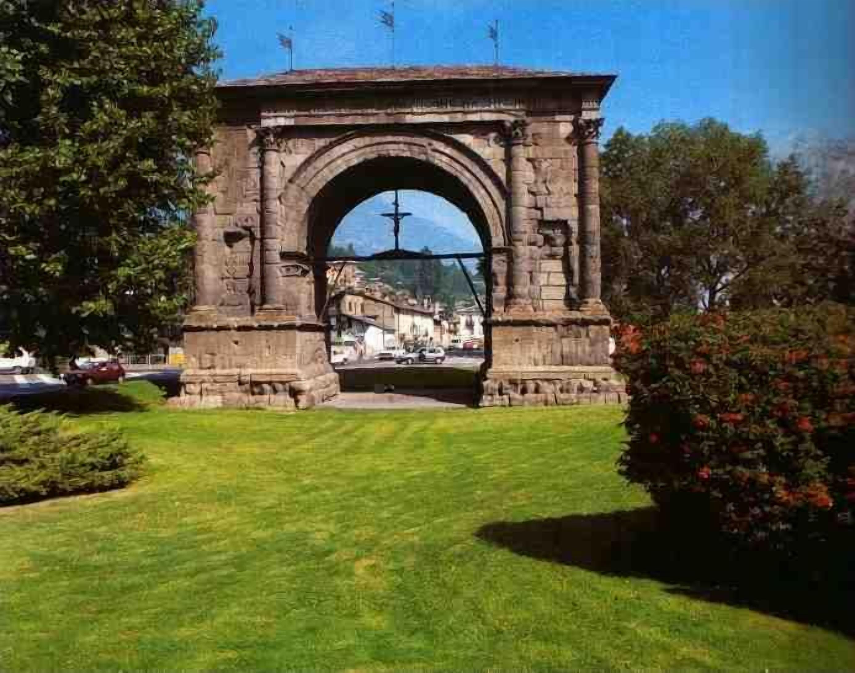 Une arche en pierre surmontée d’une statue, entourée de verdure et offrant une vue dégagée sur une ville en arrière-plan.