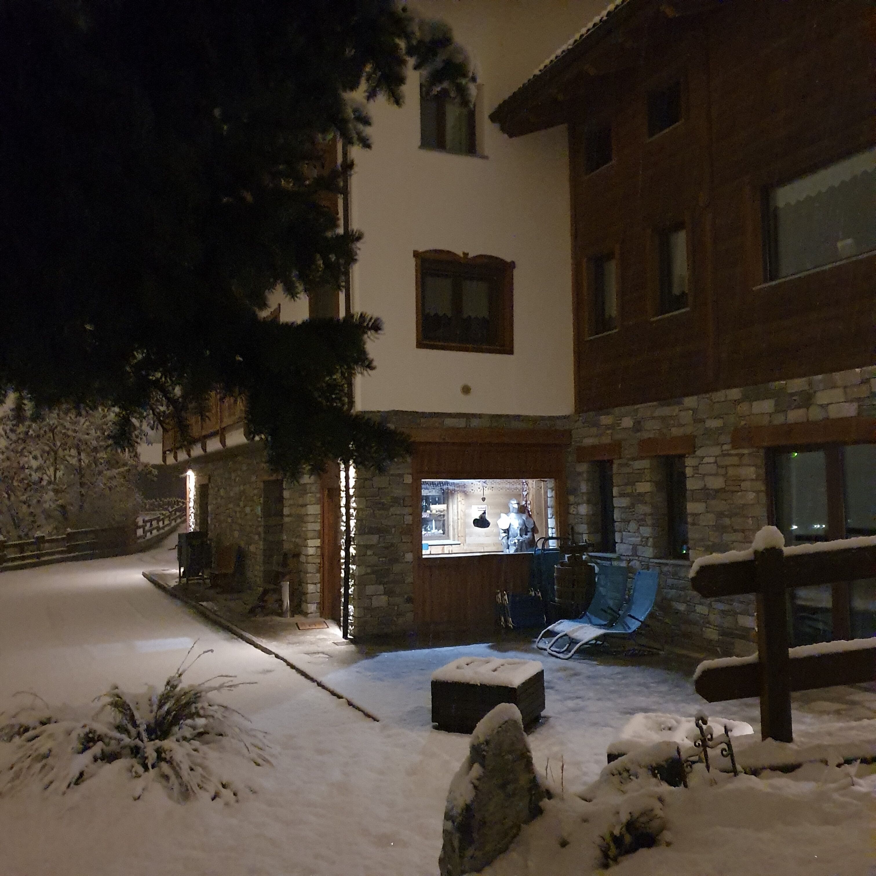 A snow-covered outdoor area with a stone building, a wooden fence, and a bench.