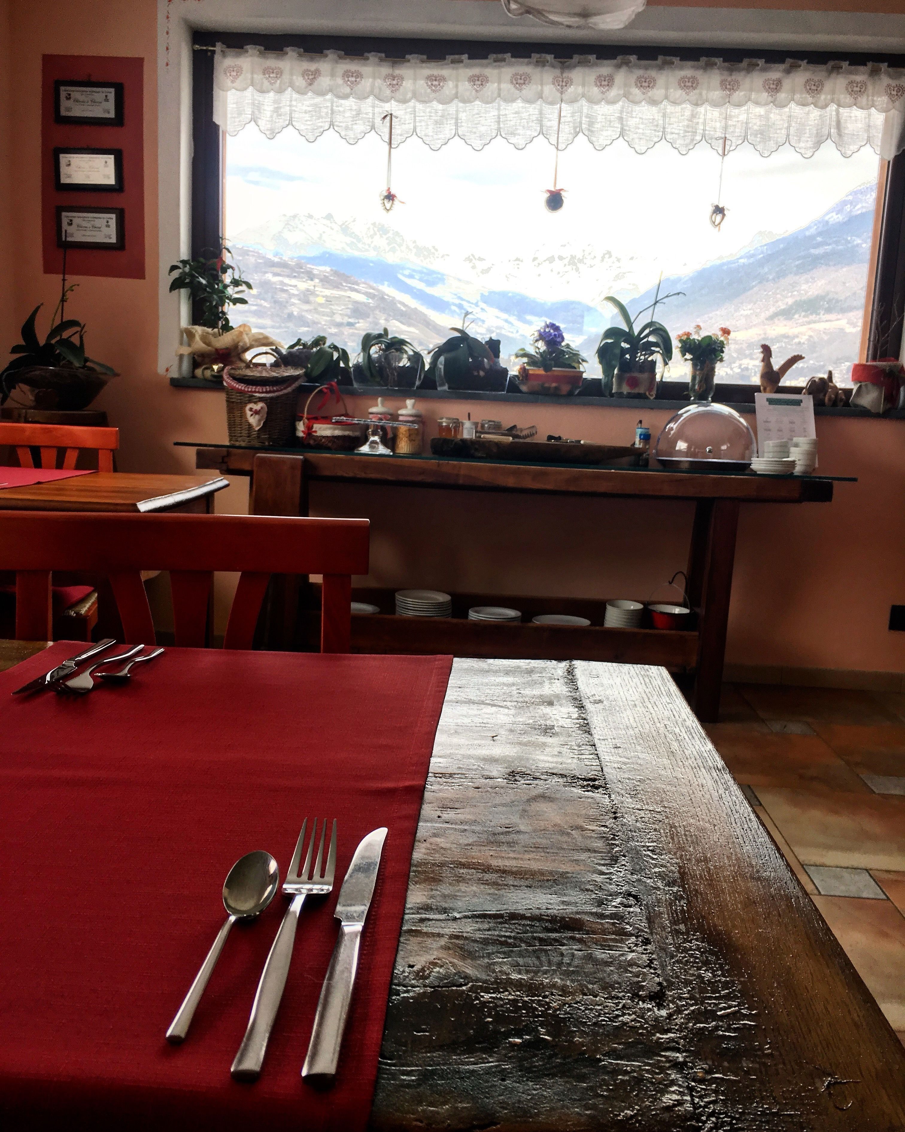 Un coin repas avec une table dressée pour deux personnes, une fenêtre donnant sur les montagnes et une étagère contenant divers objets.