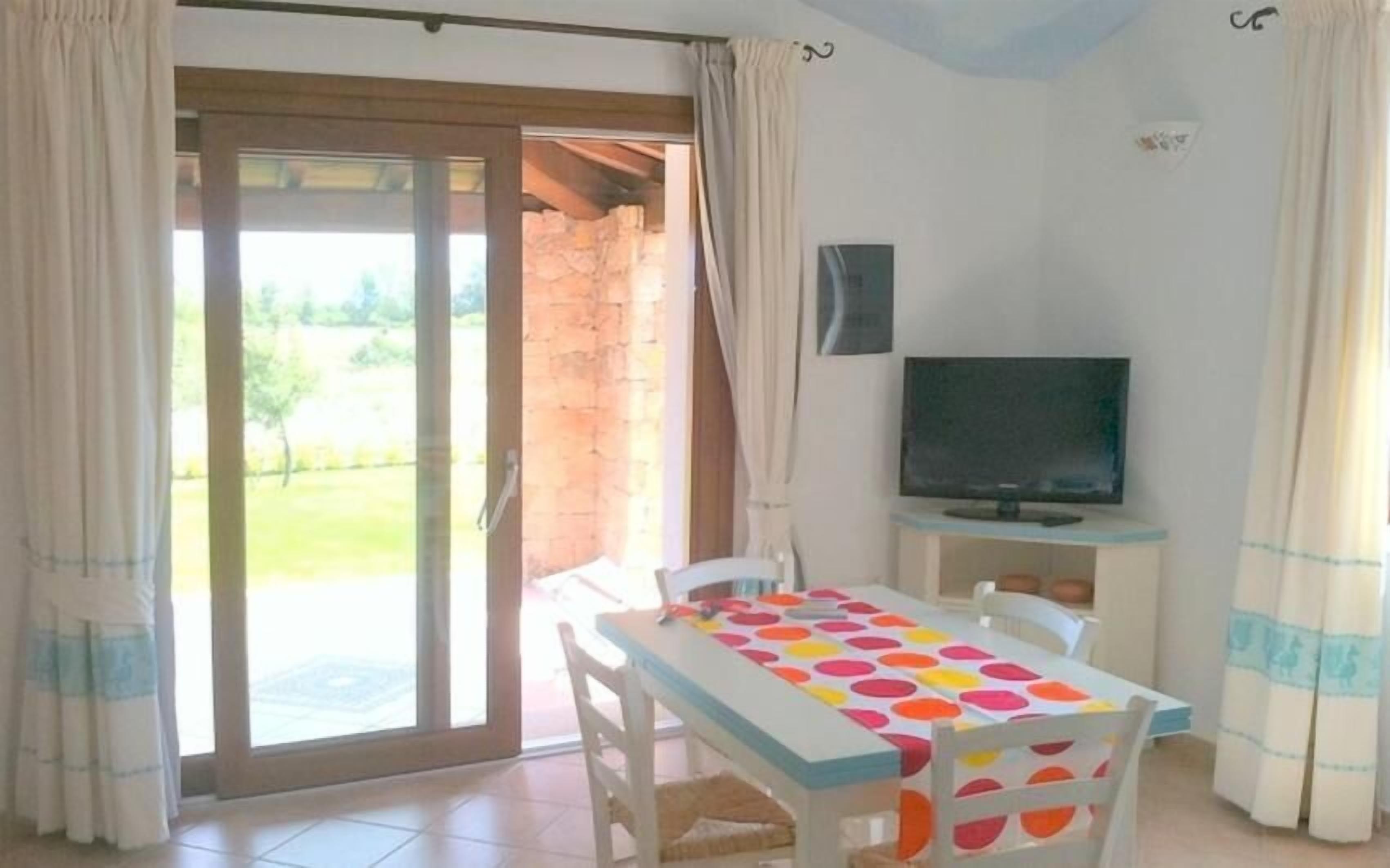 A room with a TV on a stand, a table with a colorful tablecloth, and a sliding glass door leading to an outdoor area.
