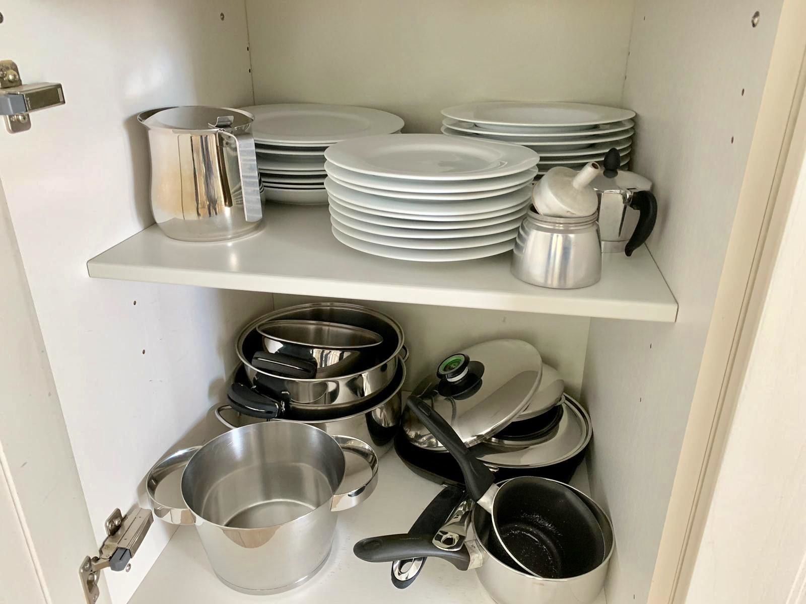 A kitchen with stainless steel cookware and white plates stacked on shelves.