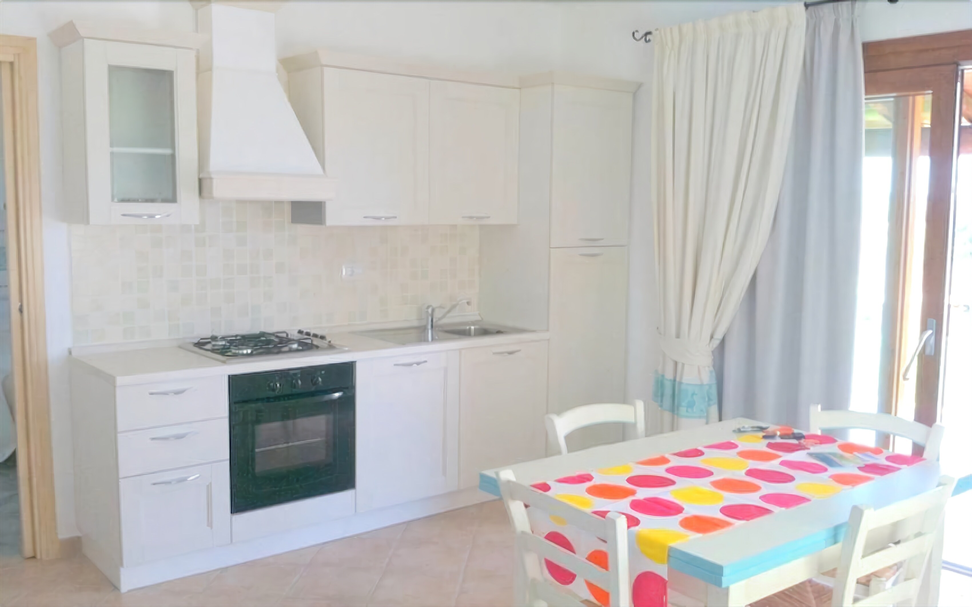 A kitchen with white cabinets, a stove, and a sink. There is a table with a colorful tablecloth and chairs.