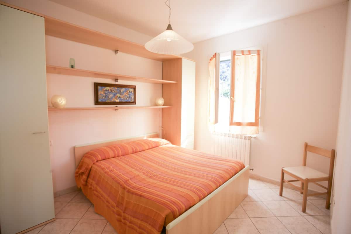 Un dormitorio con cama, una silla, estante con adornos, ventana con cortinas y lámpara colgante.