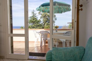 A room with a sliding glass door leading to an outdoor patio with a table and chairs, a green patterned sofa, and a view of the ocean. - Appartamenti La Bougainvillea (Marciana)