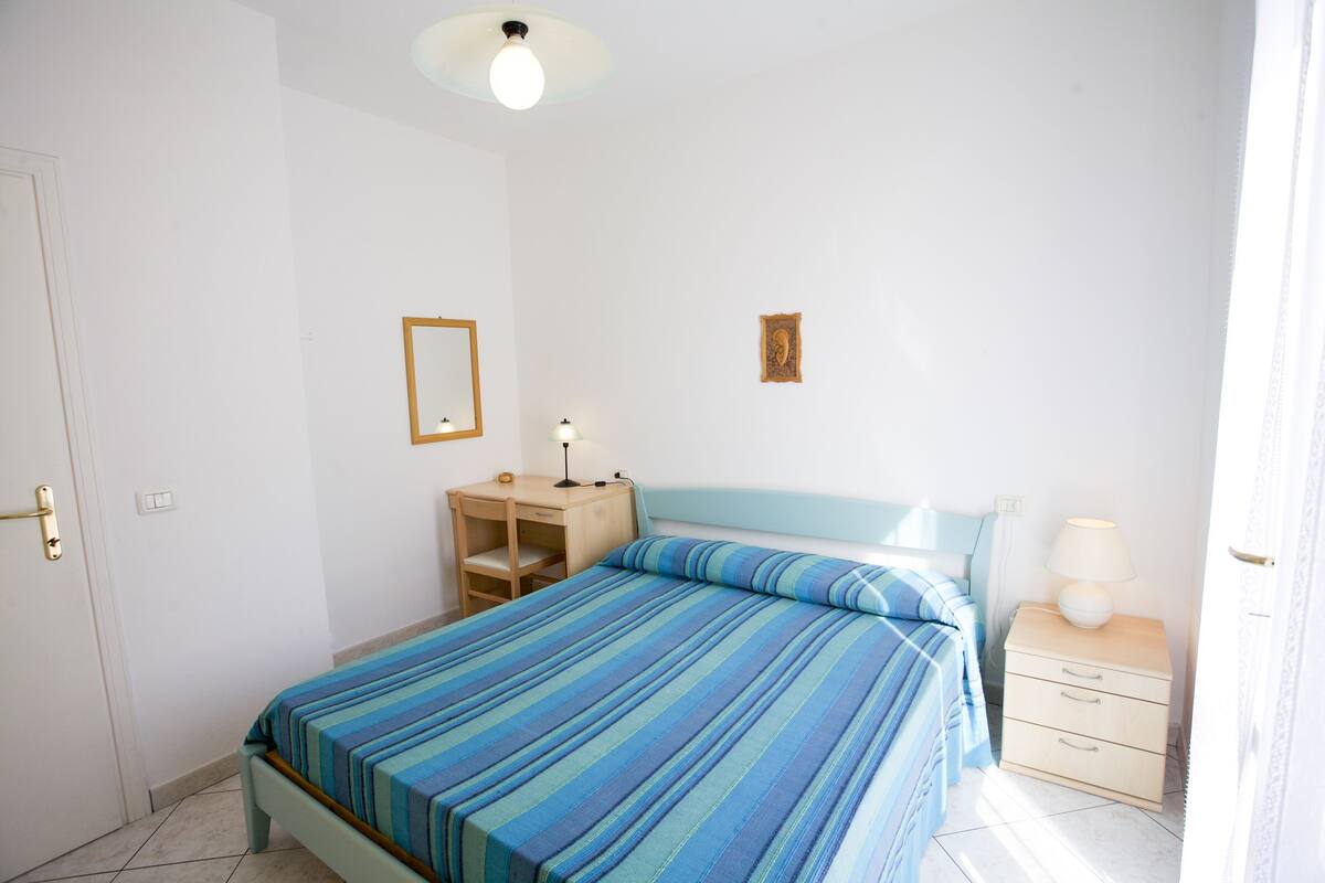 Un dormitorio con cama, mesita de noche, lámpara y un escritorio pequeño con silla.