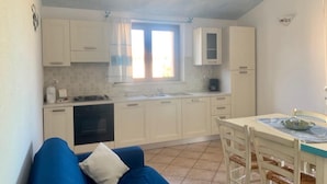 A compact kitchen with white cabinets, a dining area with a table and chairs, and a blue sofa with a pillow. - Solaria Apartment III (Budoni)