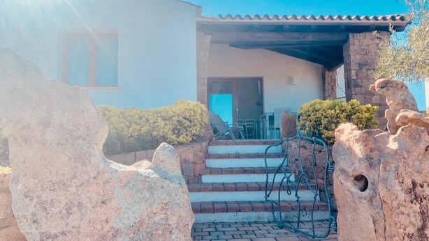 A house with a stone staircase leading to a covered entrance, surrounded by large rocks and shrubs. - Solaria Apartment III (Budoni)