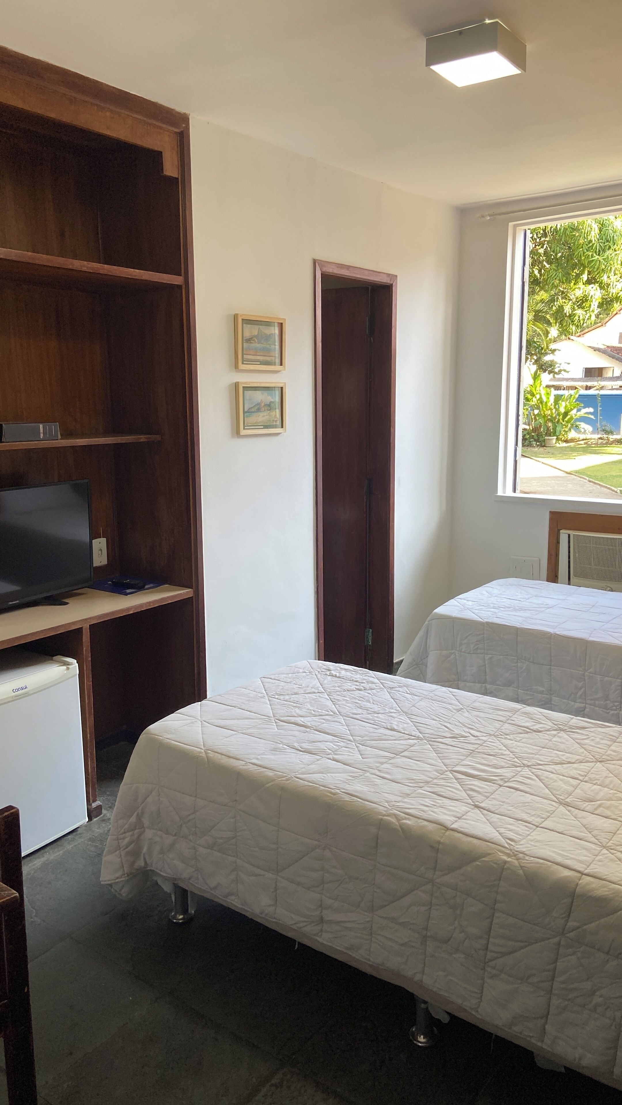 A room with two beds, a TV on a wooden cabinet, and a window with a view of greenery.