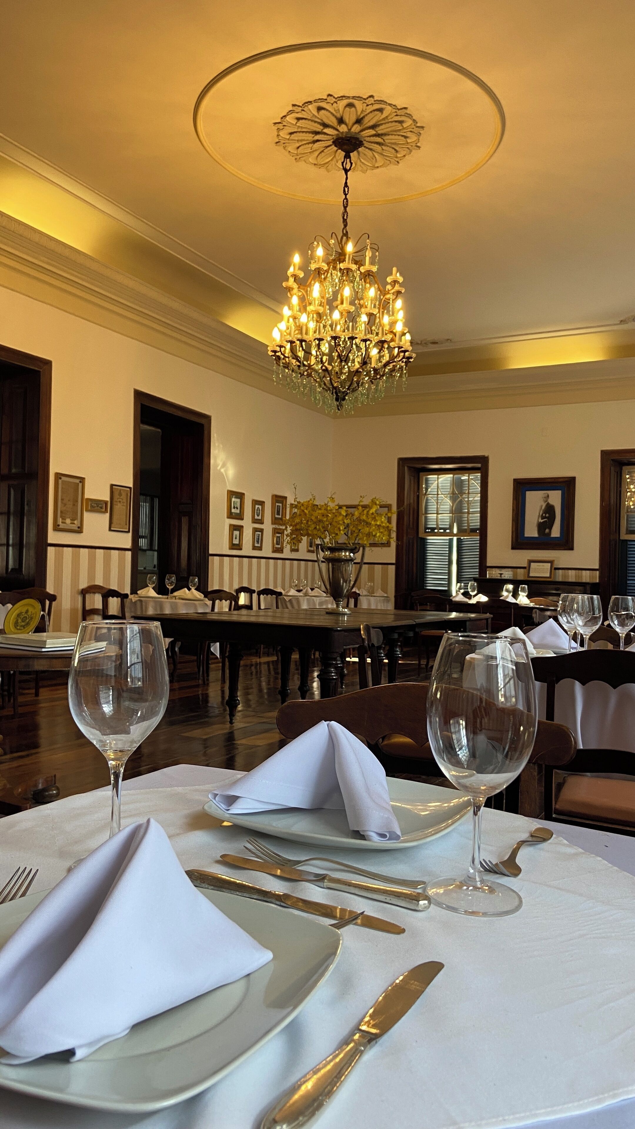 A dining area with a chandelier, tables set with white tablecloths, and a vase of yellow flowers.
