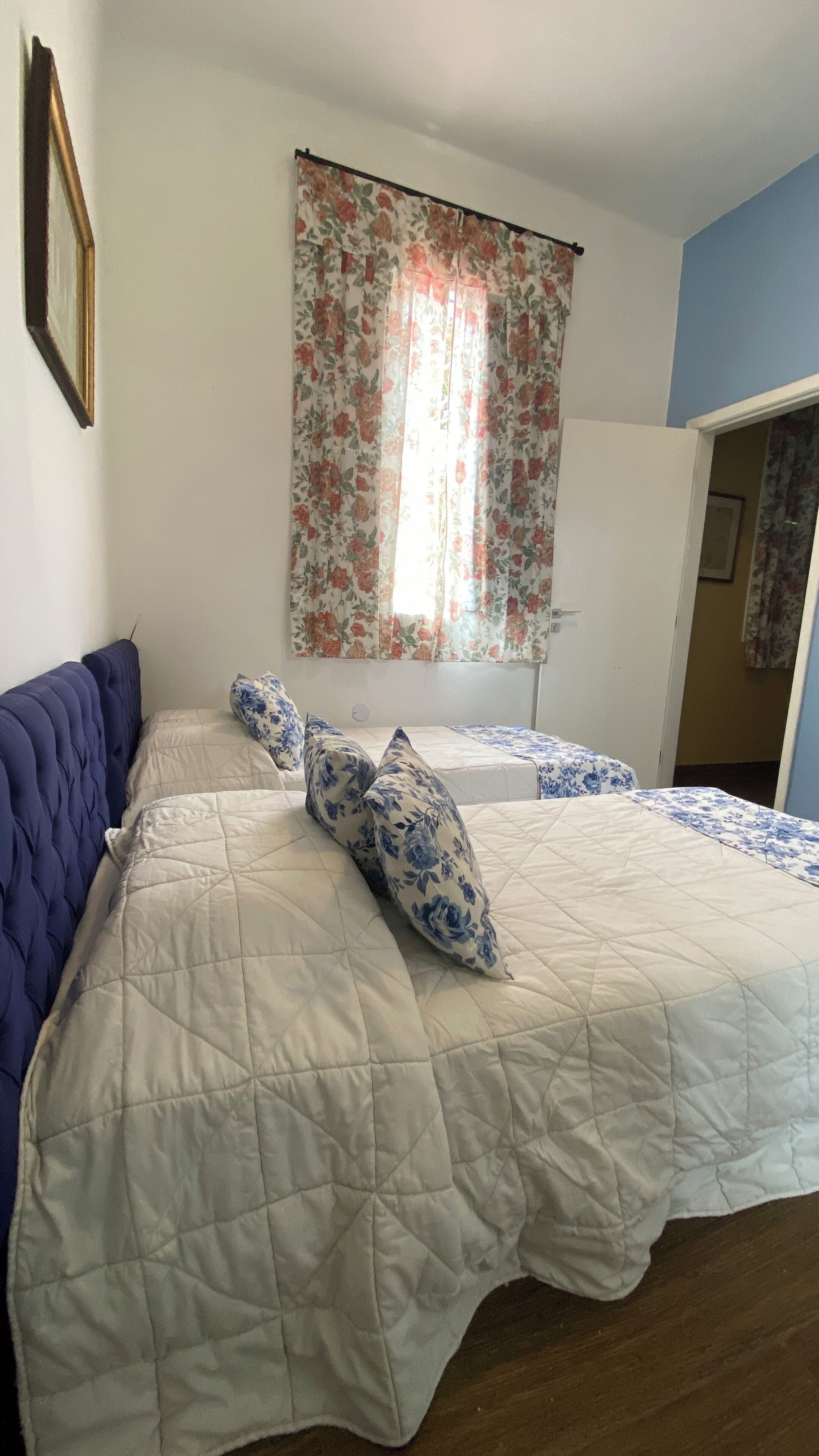A bedroom with a large bed, a blue headboard, floral curtains, and a view of a hallway.