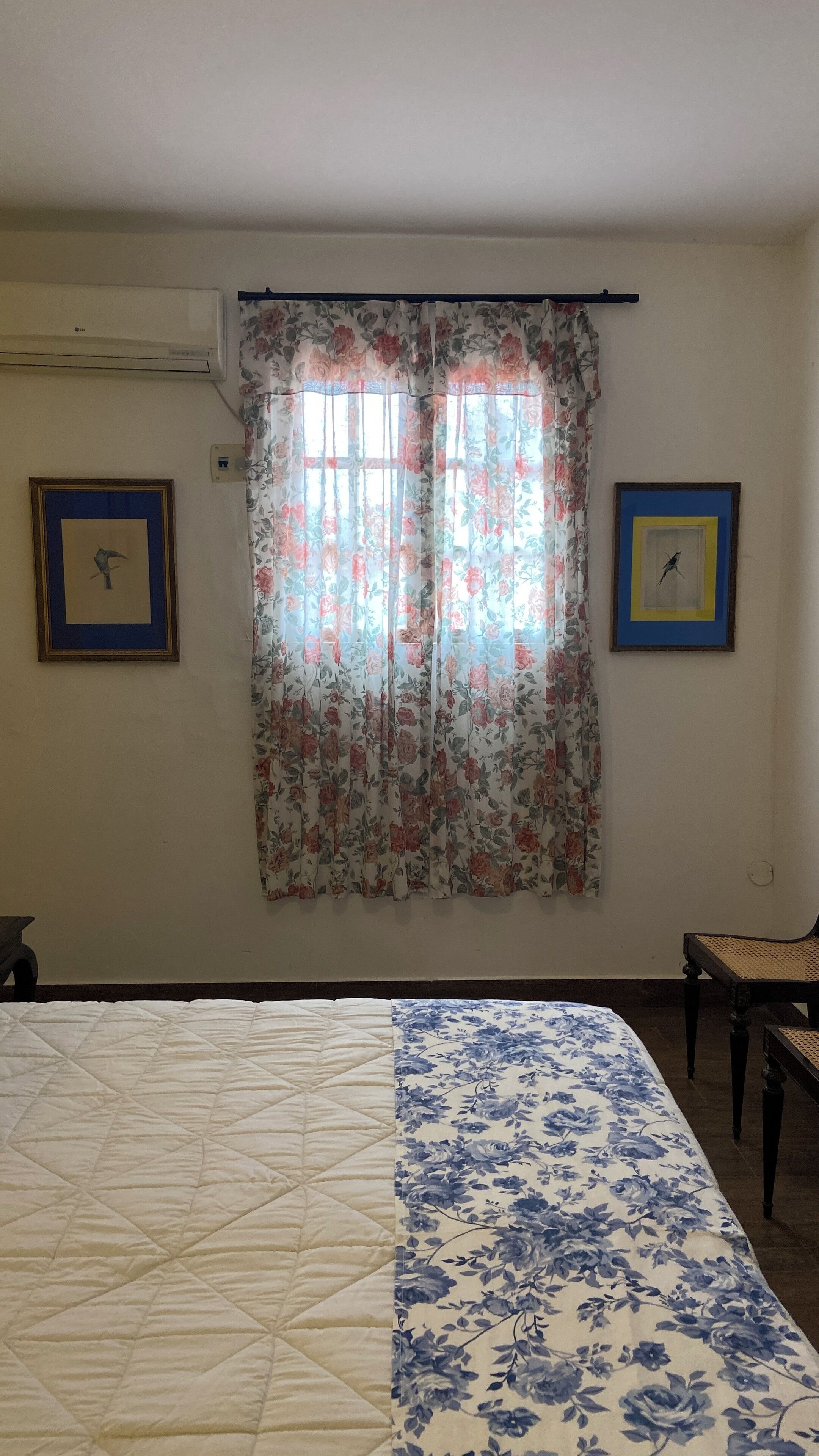 A bedroom with a bed, a chair, a window with floral curtains, and two framed pictures on the wall.