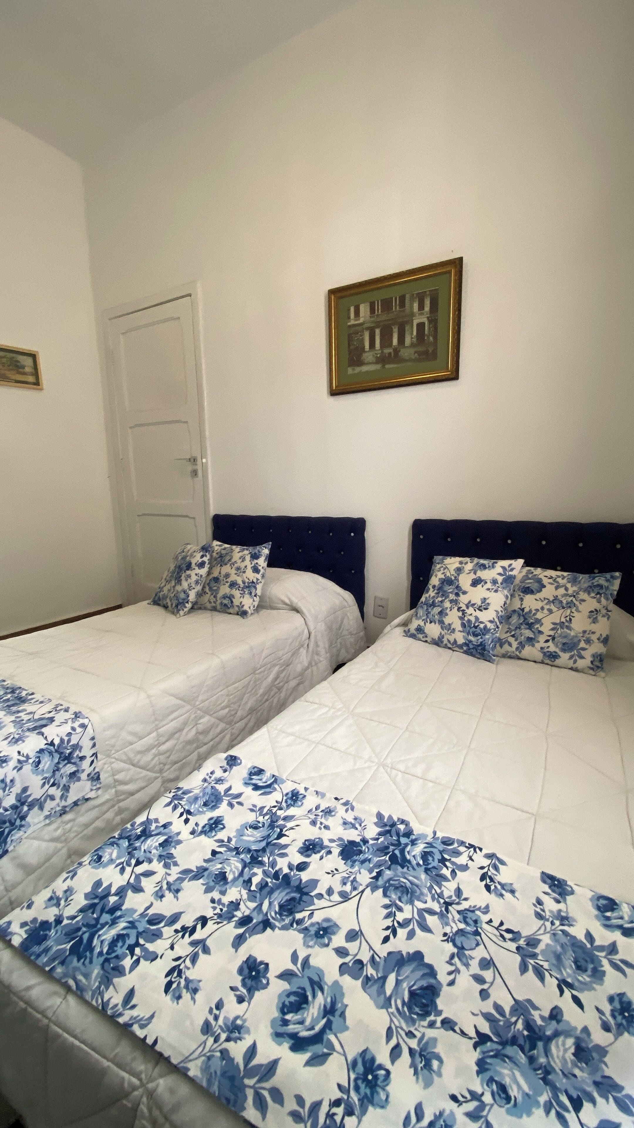 Quarto com duas camas, cada uma com cobertura e almofadas estampadas com padrão floral. Há um quadro na parede acima das camas.