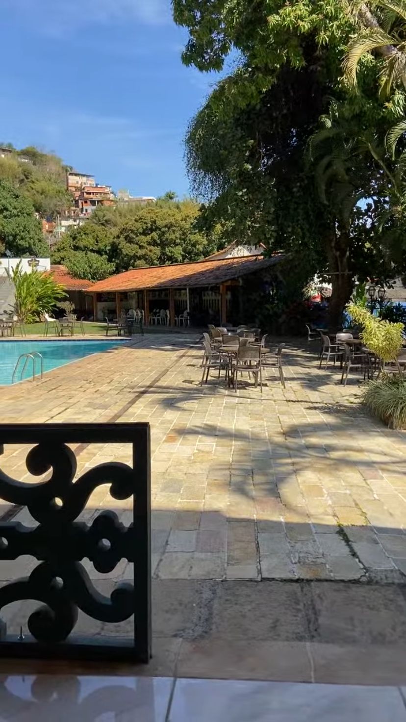 Un área junto a la piscina con suelo de piedra, mobiliario exterior y un edificio con tejado rojo.