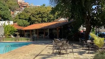 Un área junto a la piscina con suelo de piedra, mobiliario exterior y un edificio con tejado rojo.