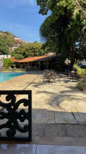 Un área junto a la piscina con suelo de piedra, mobiliario exterior y un edificio con tejado rojo.