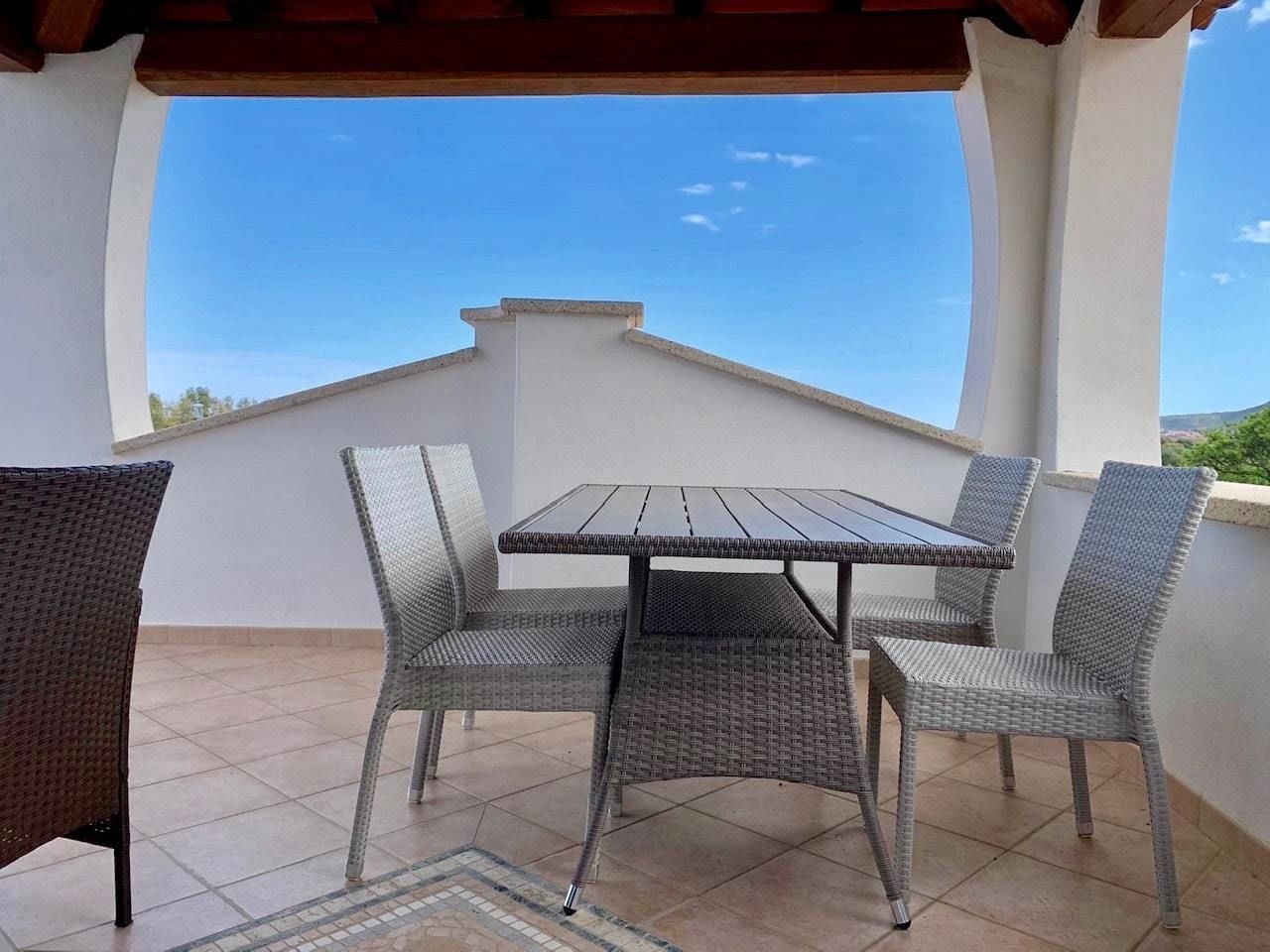 Een terras buiten met een tafel en vier rieten stoelen, een betegelde vloer en een helderblauwe lucht.