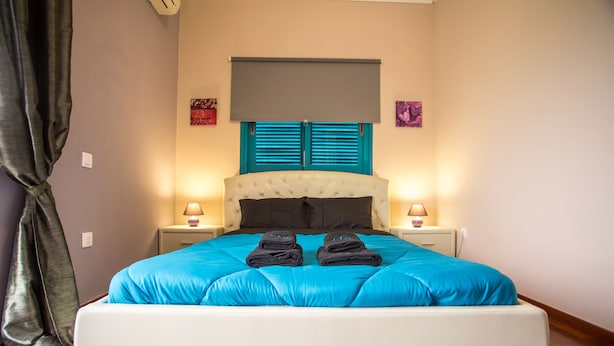 Una cama bien hecha con una colcha azul y dos toallas dobladas, un cabecero blanco y dos lámparas de noche en mesitas.