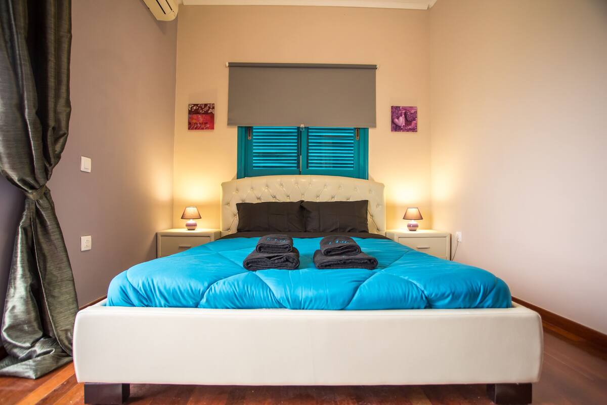 A neatly made bed with a blue bedspread and two folded towels, a white headboard, and two bedside lamps on nightstands.