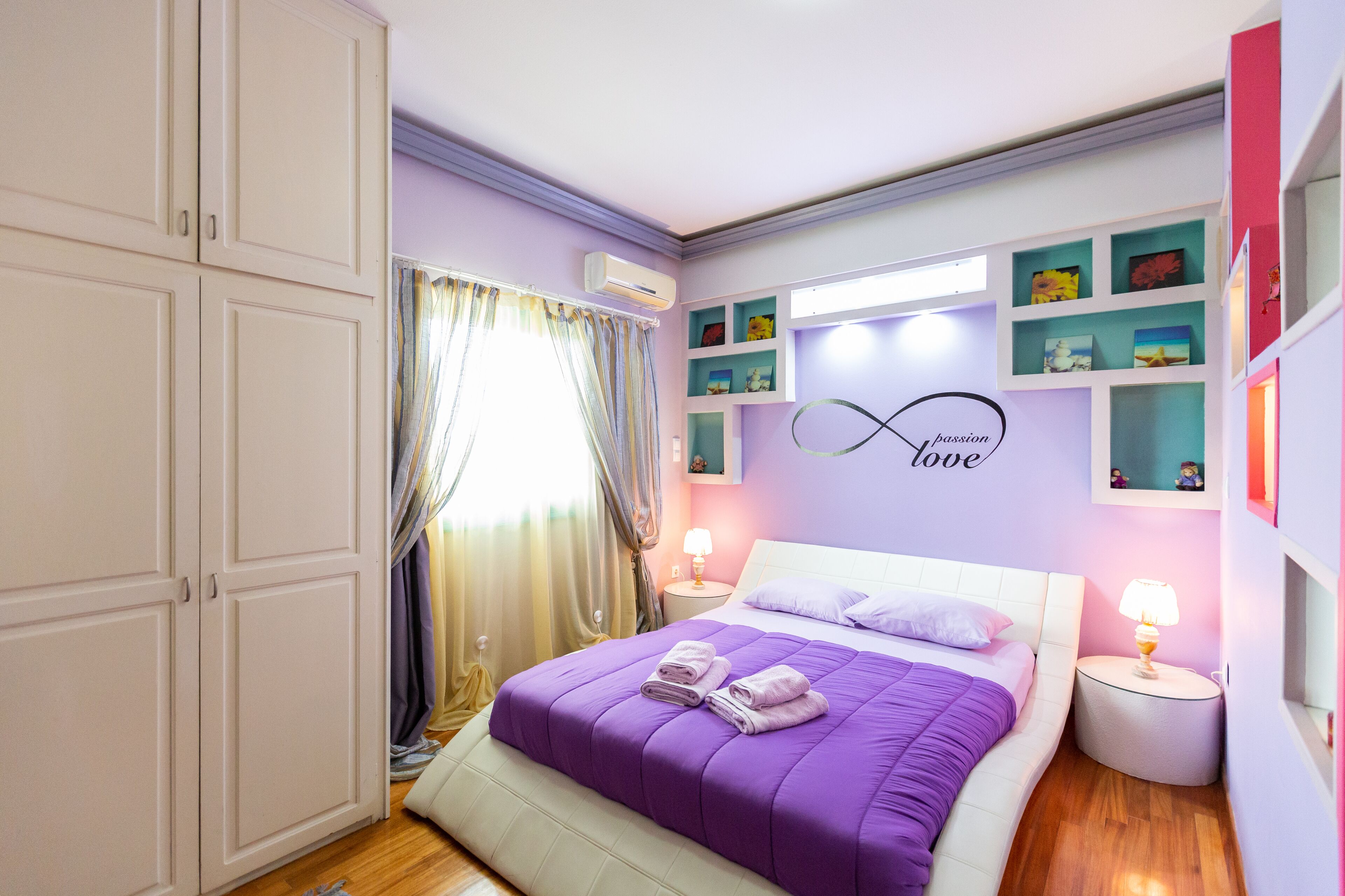 Une chambre à coucher avec une couvre-lit violette, une tête de lit blanche et un mur orné d’étagères où sont exposées des œuvres d’art.
