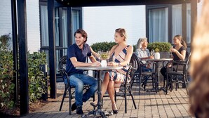 Une terrasse extérieure avec des tables et des chaises, entourée de verdure et bordée d’un bâtiment doté de grandes fenêtres.