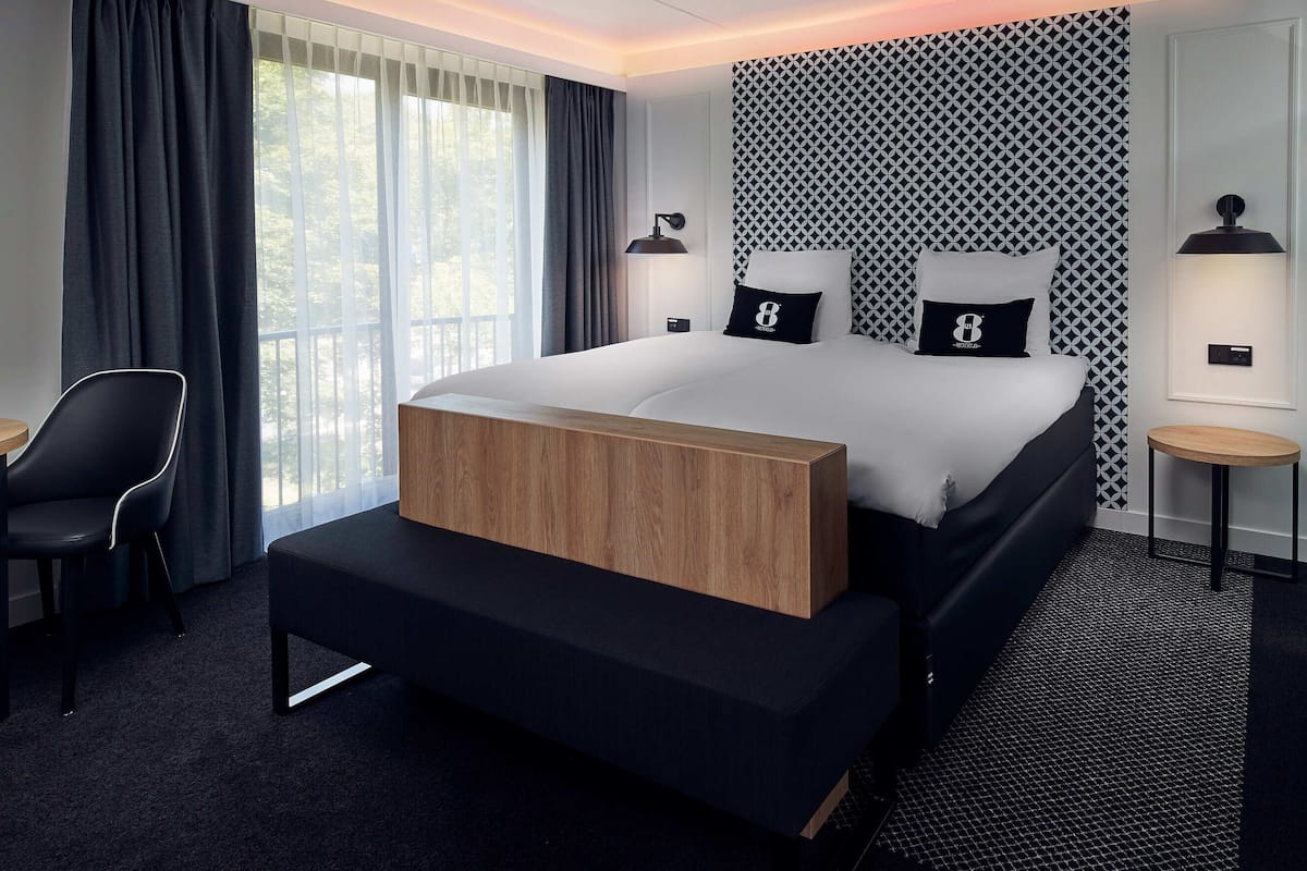 Een moderne hotelkamer met een groot bed, twee nachtkastjes met lampen, een houten nachtkastje, een zwarte stoel, en uitzicht op groen door het raam.