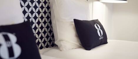 A neatly made bed with white bedding and two pillows, one with a logo and another plain white. A patterned wall and a modern lamp are visible in the background.