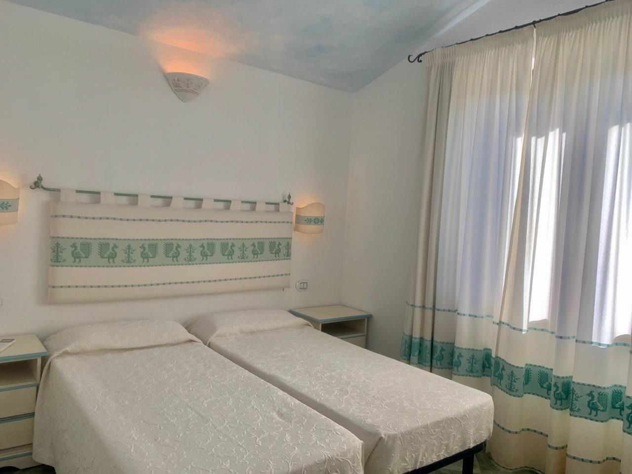Una habitación con dos camas individuales, una mesita de noche, una lámpara de pared y un ventanal con cortinas.