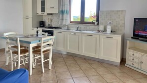 A compact kitchen with white cabinets, a dining table with chairs, a TV on a stand, and a window with curtains. - Solaria Apartment VIII (Budoni)
