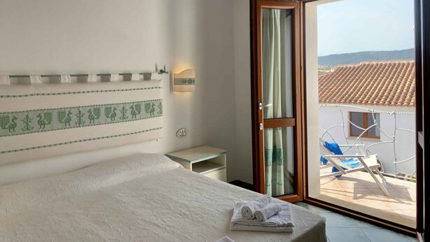 A hotel room with a bed, a nightstand, a wall-mounted light, a sliding glass door to a balcony, and a view of a tiled roof outside. - Solaria Apartment VIII (Budoni)