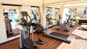 A covered outdoor gym with various exercise machines, a wooden deck, and a view of a swimming pool and buildings in the background. - Hotel Conchiglia (Cervia)