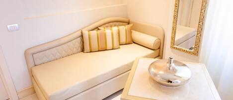 Un dormitorio moderno y minimalista con un sofá color beige, una mesita con un objeto plateado y un espejo con marco dorado.