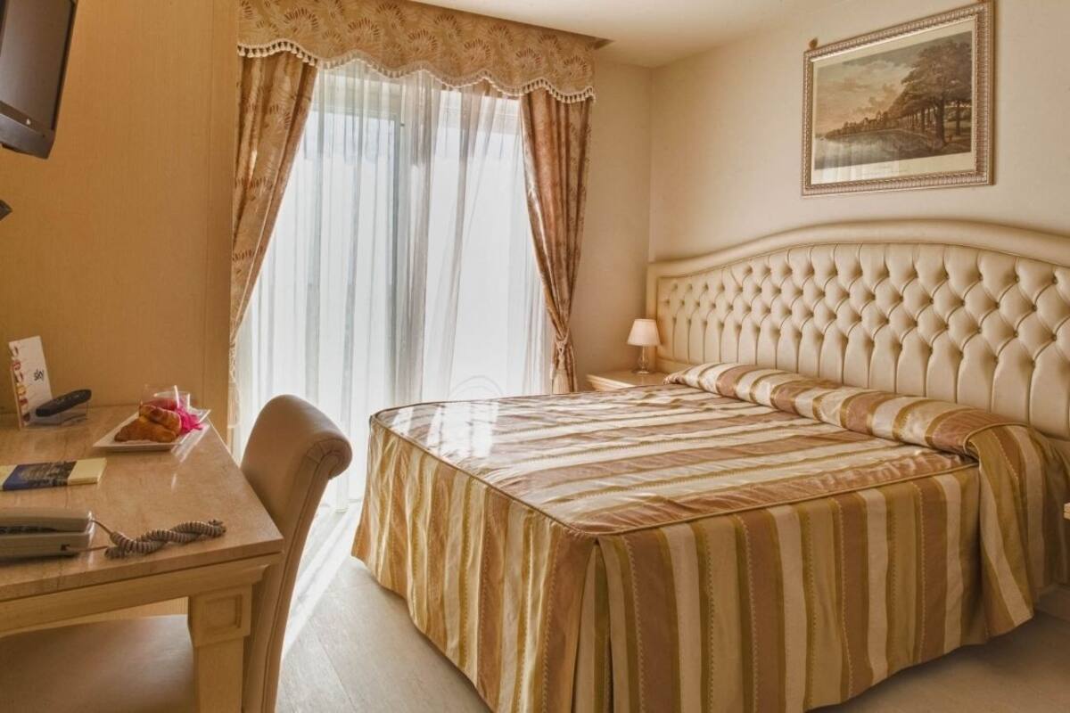 A hotel room with a large bed, a desk with a telephone and a lamp, and a window with curtains.