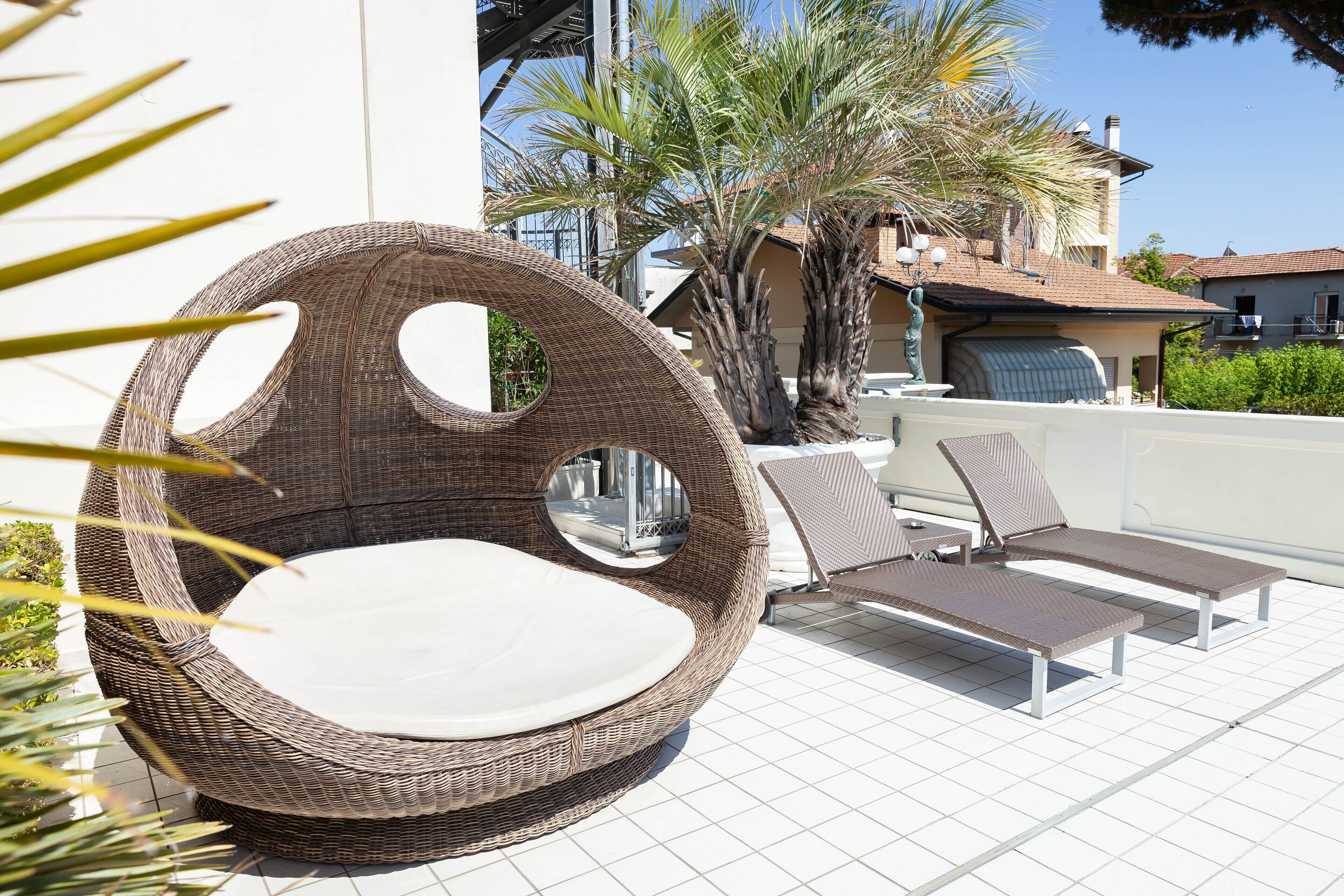 Une chaise en osier avec un miroir rond, deux fauteuils de relaxation et un palmier sur une terrasse carrelée.