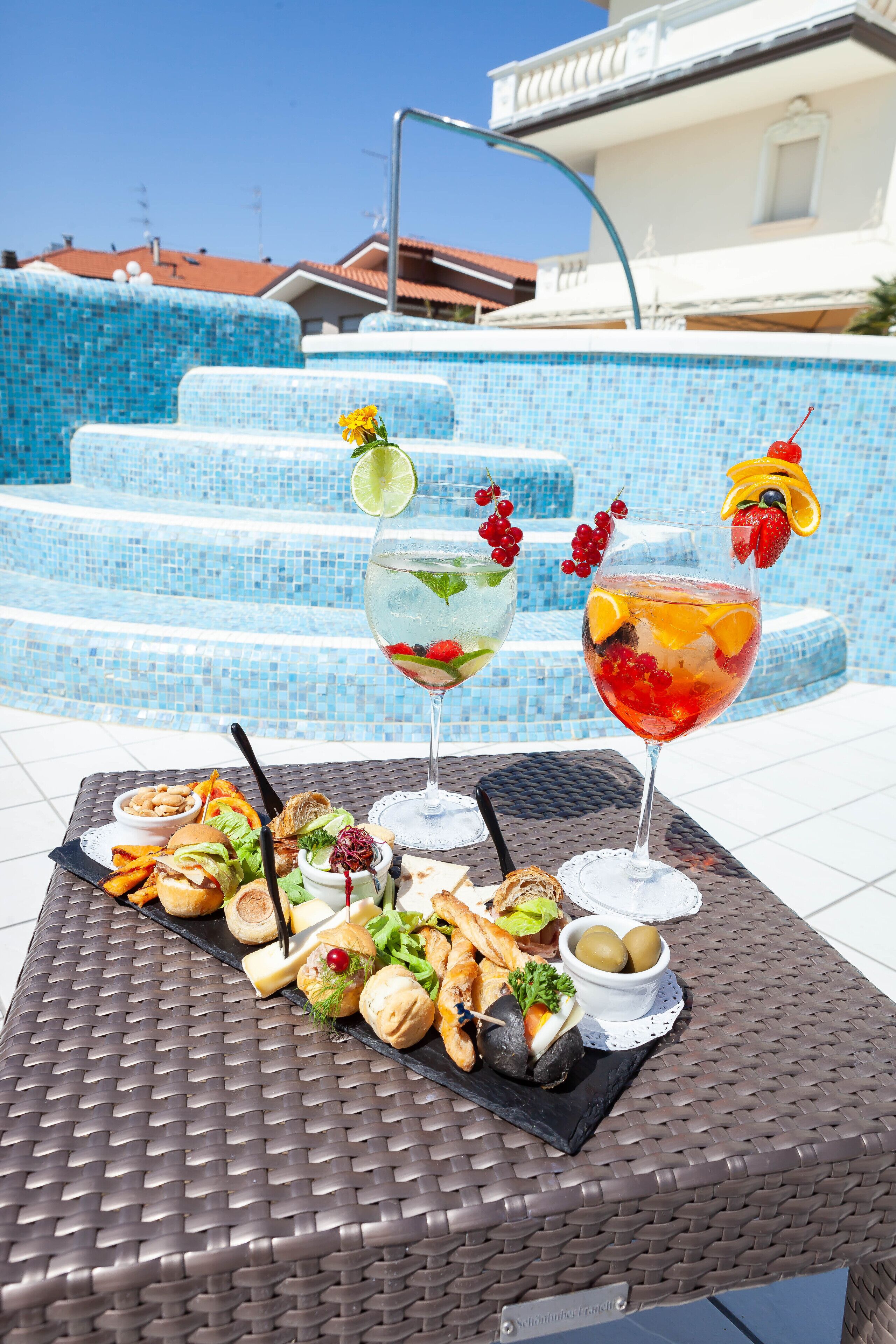 A table with a variety of food and drinks, a pool with blue mosaic tiles, and a building with a red roof in the background.