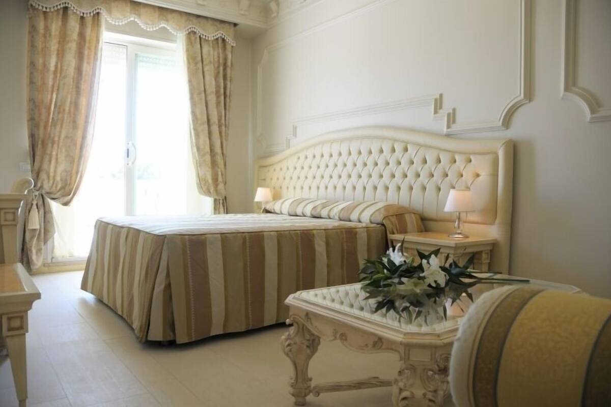 A bedroom with two beds, a large window with curtains, a bedside table with a lamp, and a decorative vase with flowers.