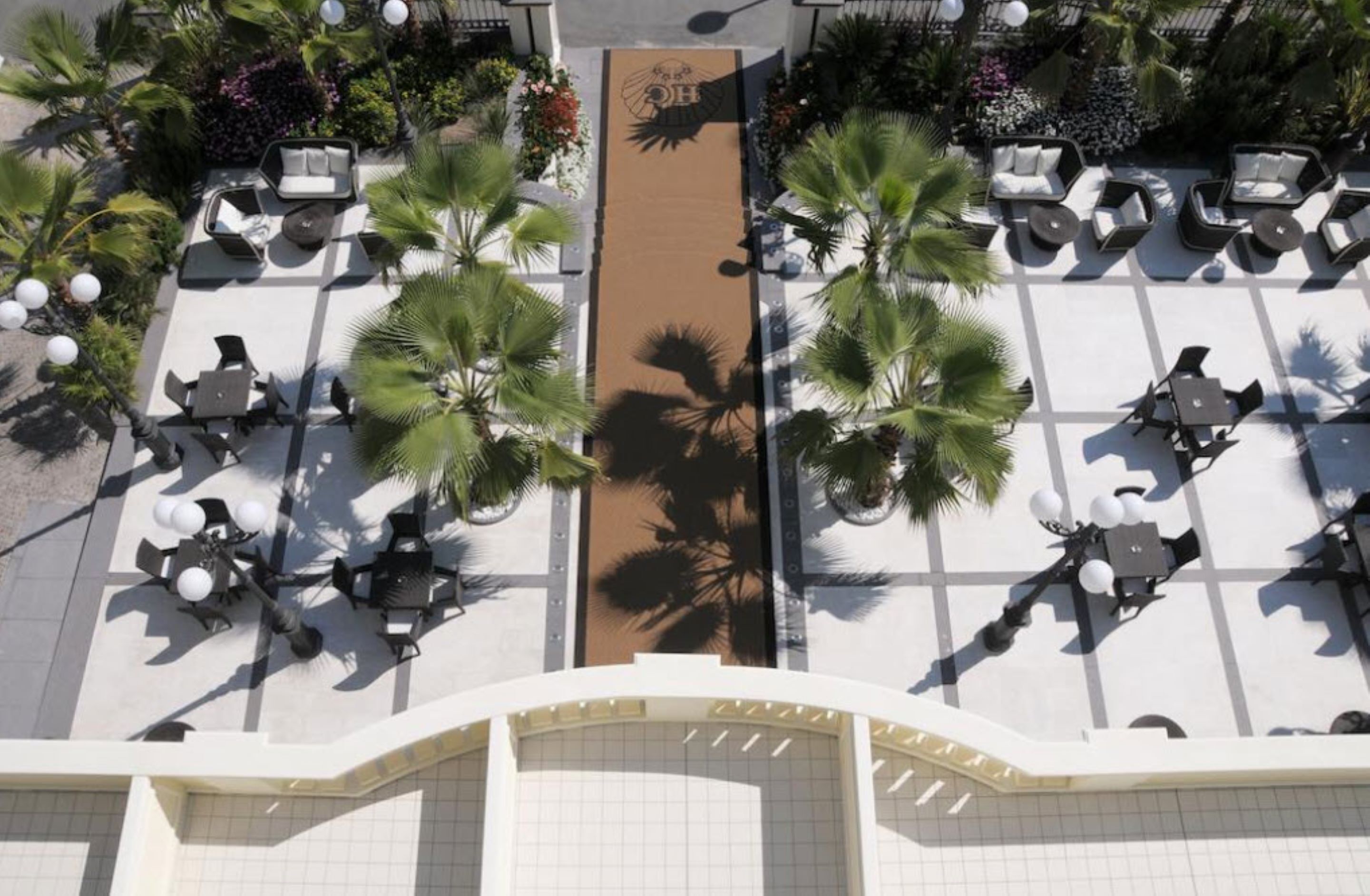 Vista aerea di un cortile alberghiero con aree salotto all'aperto, palme e un percorso centrale.
