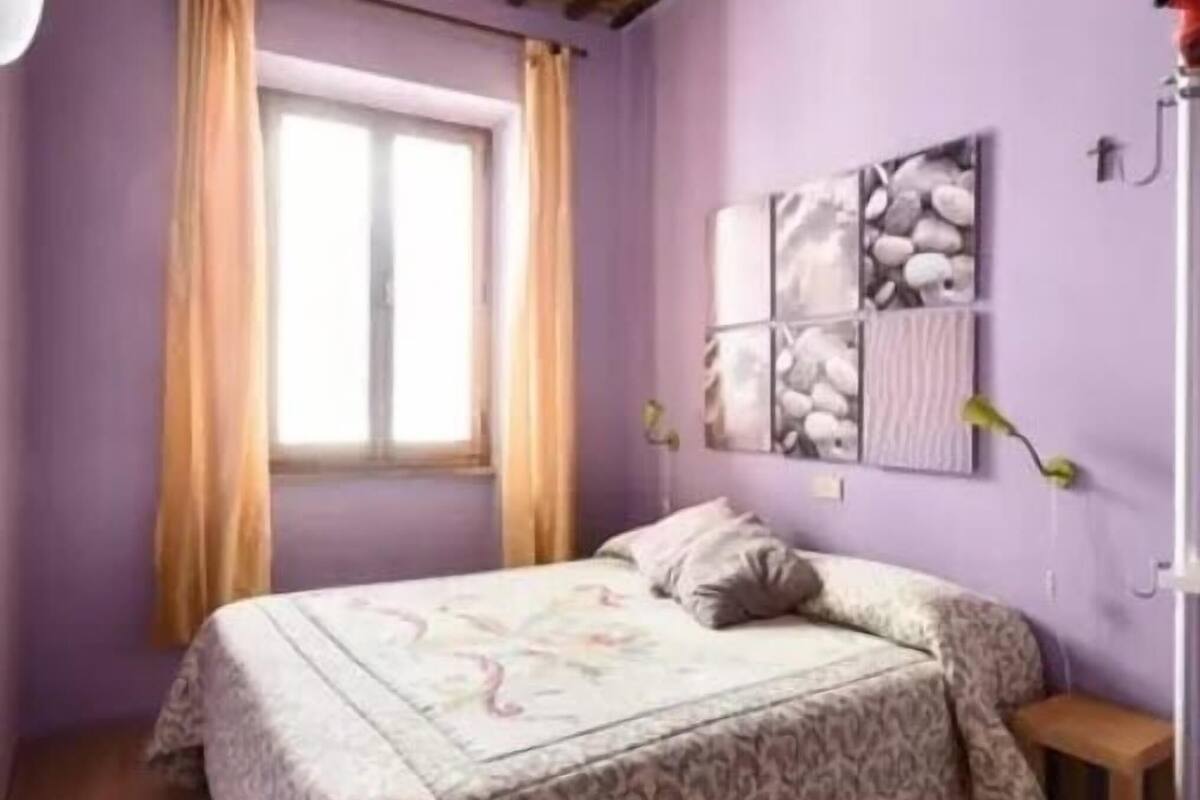 Un dormitorio con una cama, una ventana con cortinas, una obra de arte en la pared y una mesita de noche pequeña.