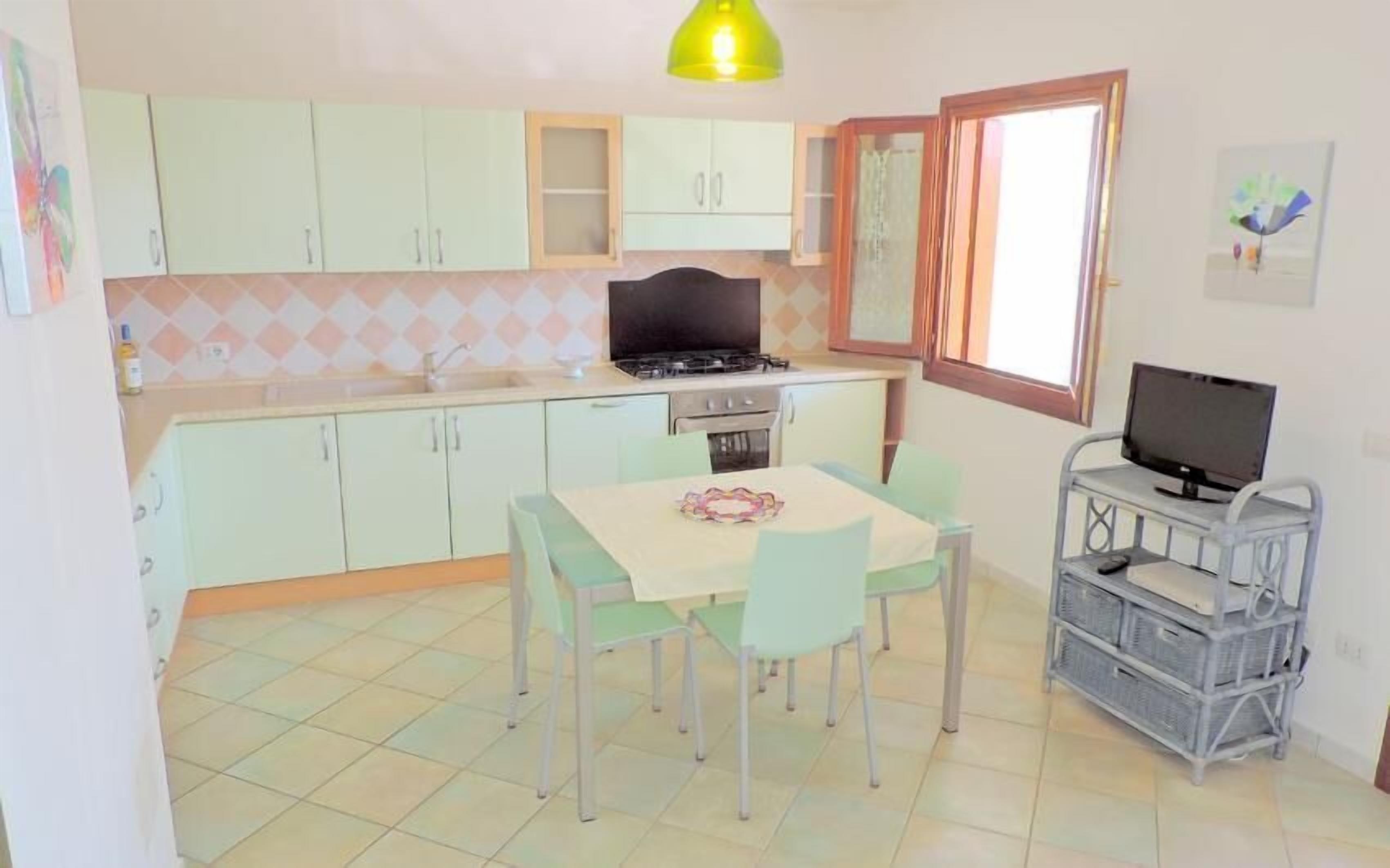 Cocina compacta con gabinetes verde claro, estufa a gas, pileta y comedor con mesa y sillas. Un televisor sobre un mueble se encuentra a la derecha.