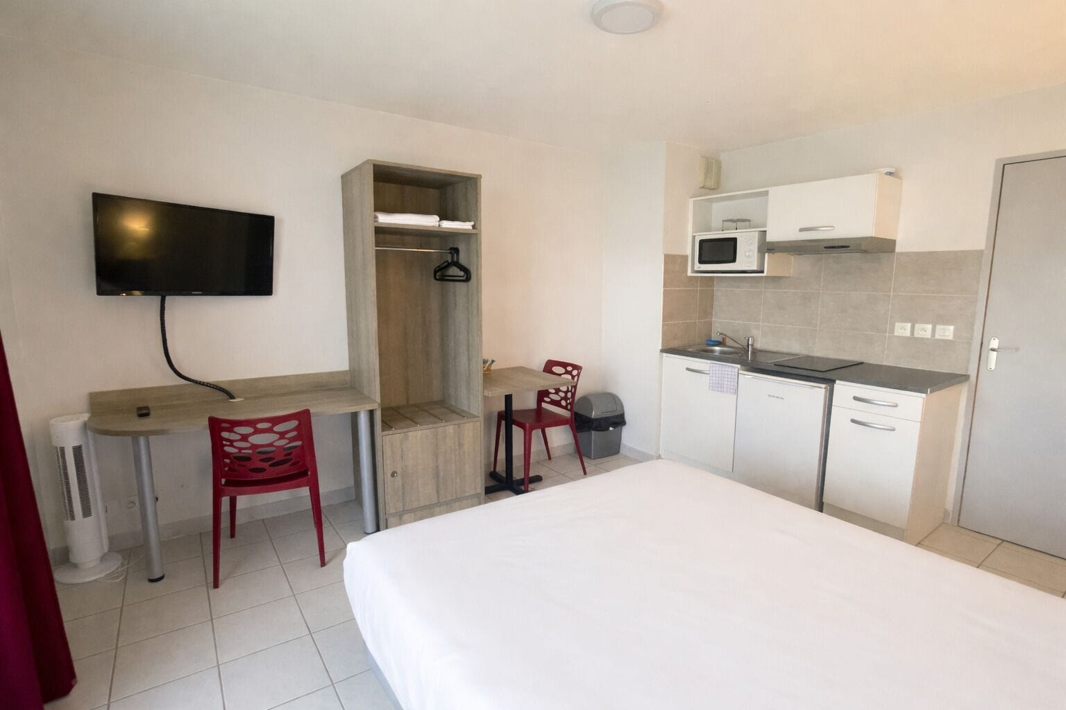 Une chambre d’hôtel de taille réduite, comprenant un lit, un bureau, une chaise, une télévision, une armoire et une kitchenette.