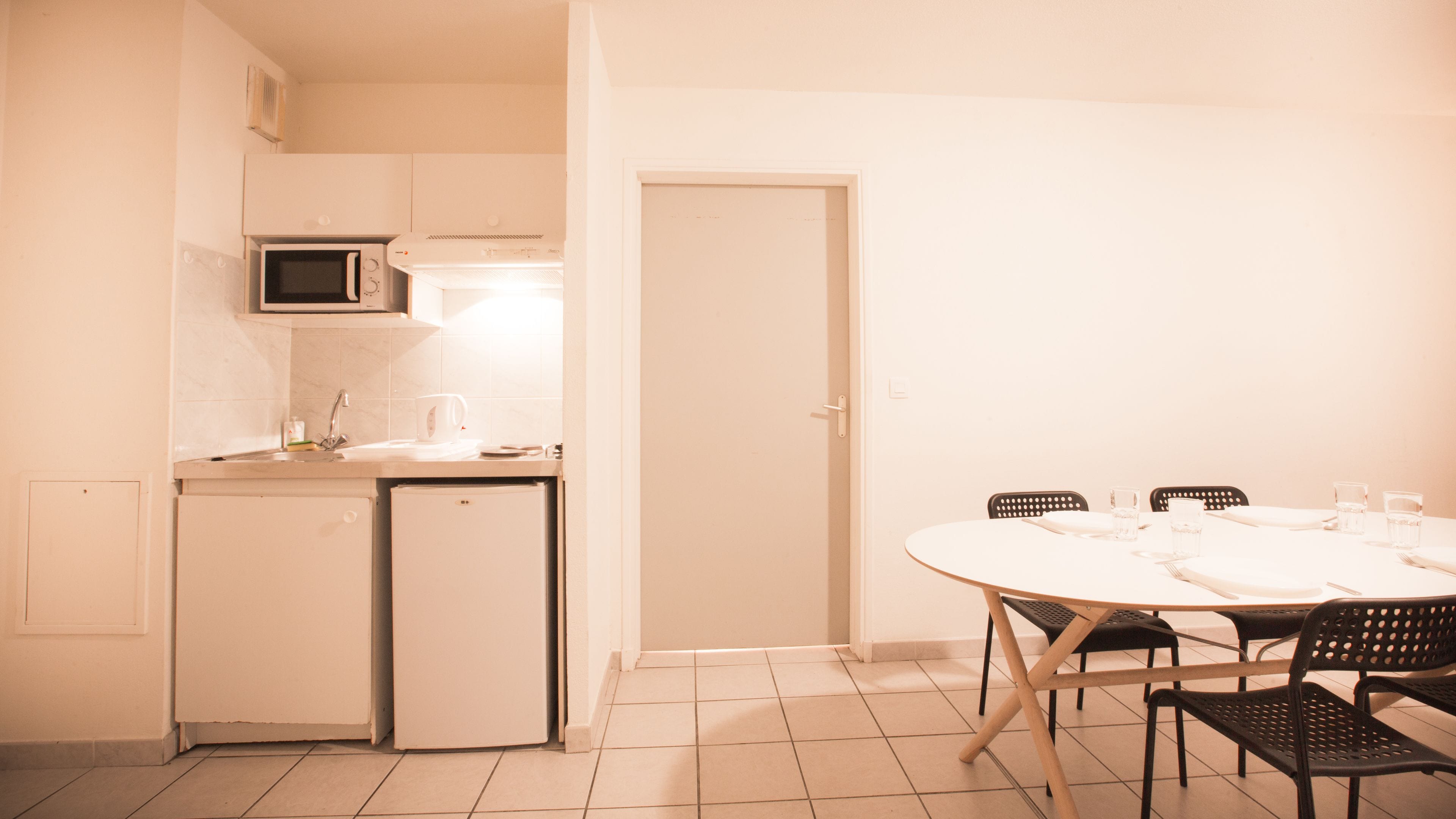 Cocina compacta con microondas, cafetera y refrigerador pequeño. Hay una mesa de comedor con sillas y una puerta que da a otra habitación.