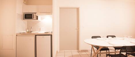 Cocina compacta con microondas, cafetera y refrigerador pequeño. Hay una mesa de comedor con sillas y una puerta que da a otra habitación.