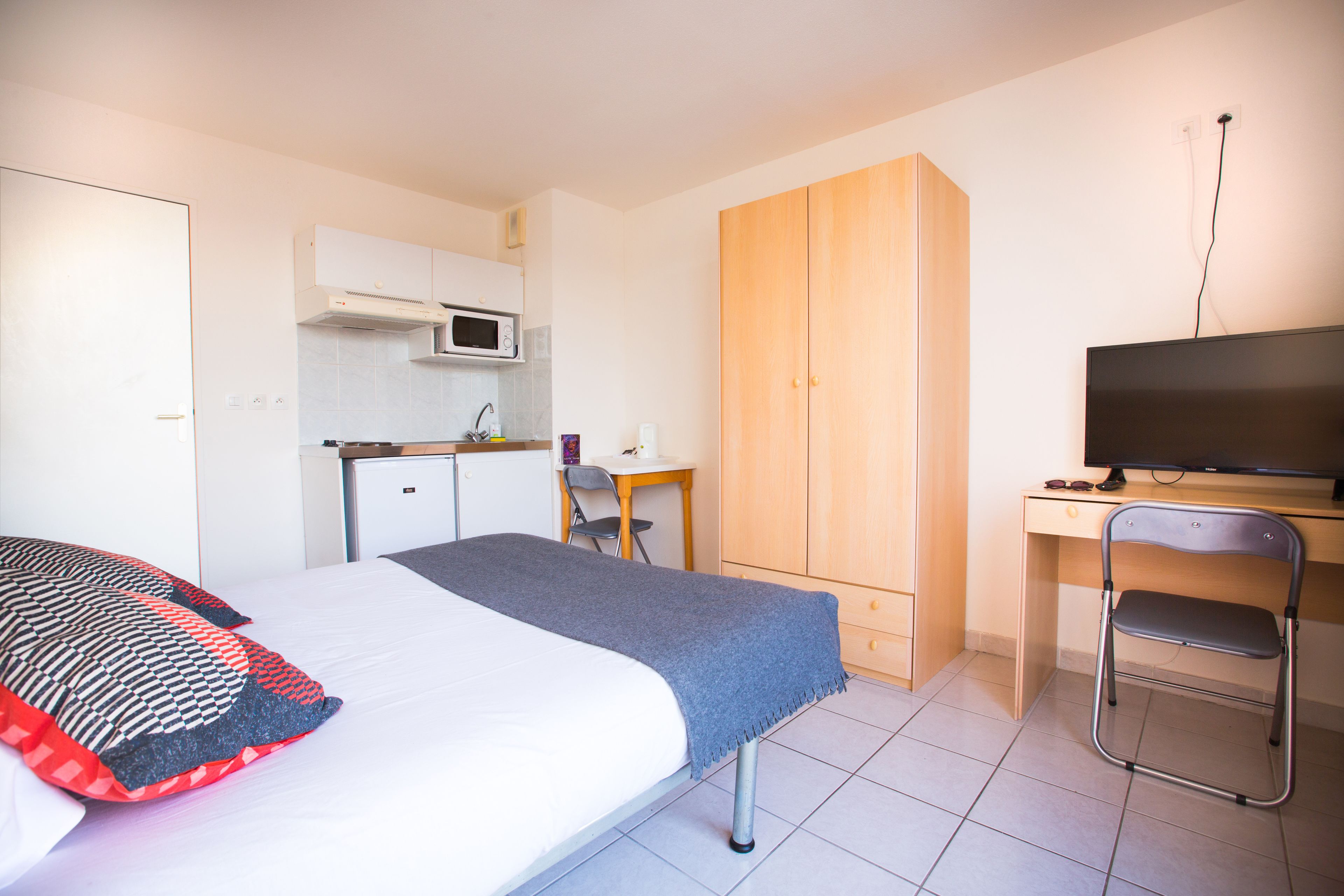 A room with a bed, a desk, a chair, a TV, and a kitchenette.