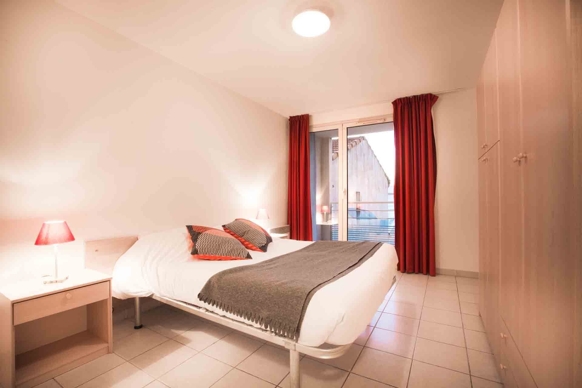 Une chambre à coucher avec un lit, une table de chevet, une lampe et une vue sur l’extérieur par une fenêtre ornée de rideaux rouges.