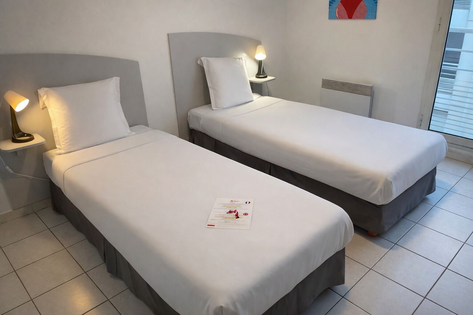 A hotel room with two single beds, each with a bedside table and lamp.