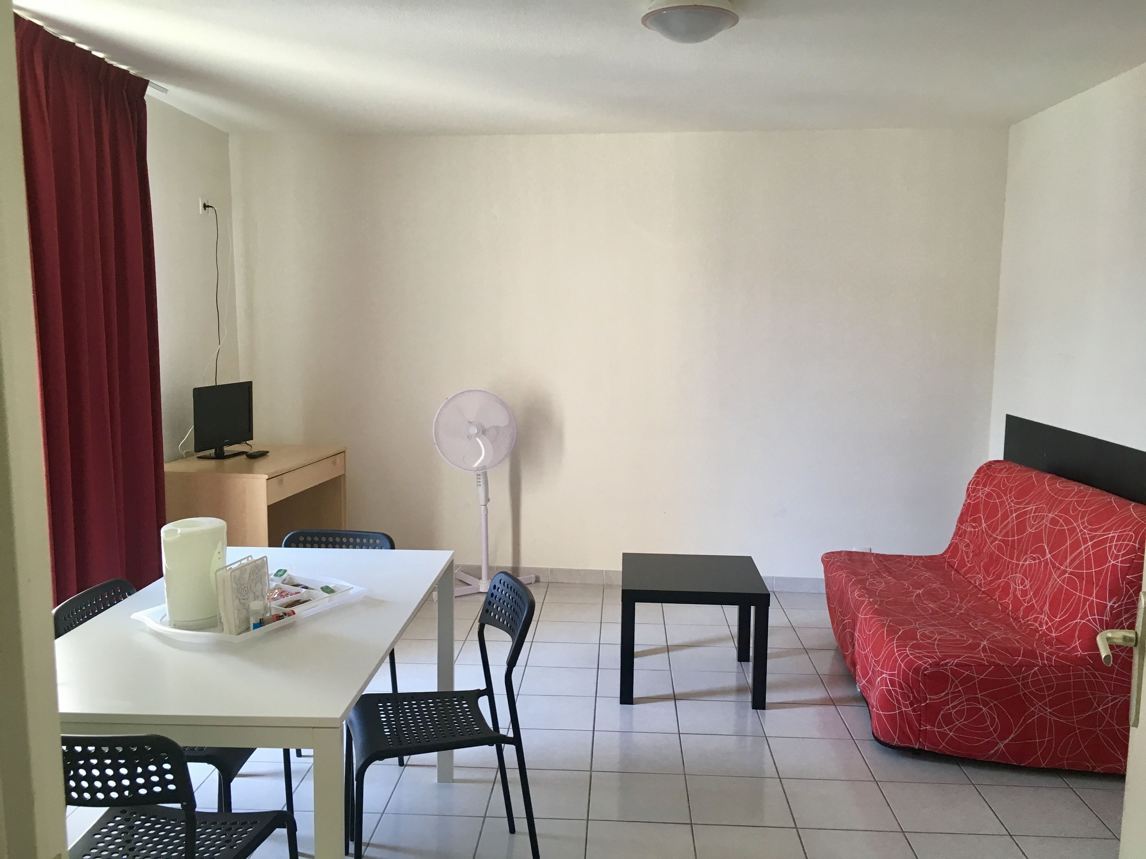 A small, well-lit room with a white table, a red sofa, a black chair, a small black table, and a standing fan.