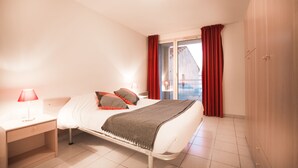 Une chambre à coucher avec un lit, une table de chevet, une lampe et une vue sur l’extérieur par une fenêtre ornée de rideaux rouges.