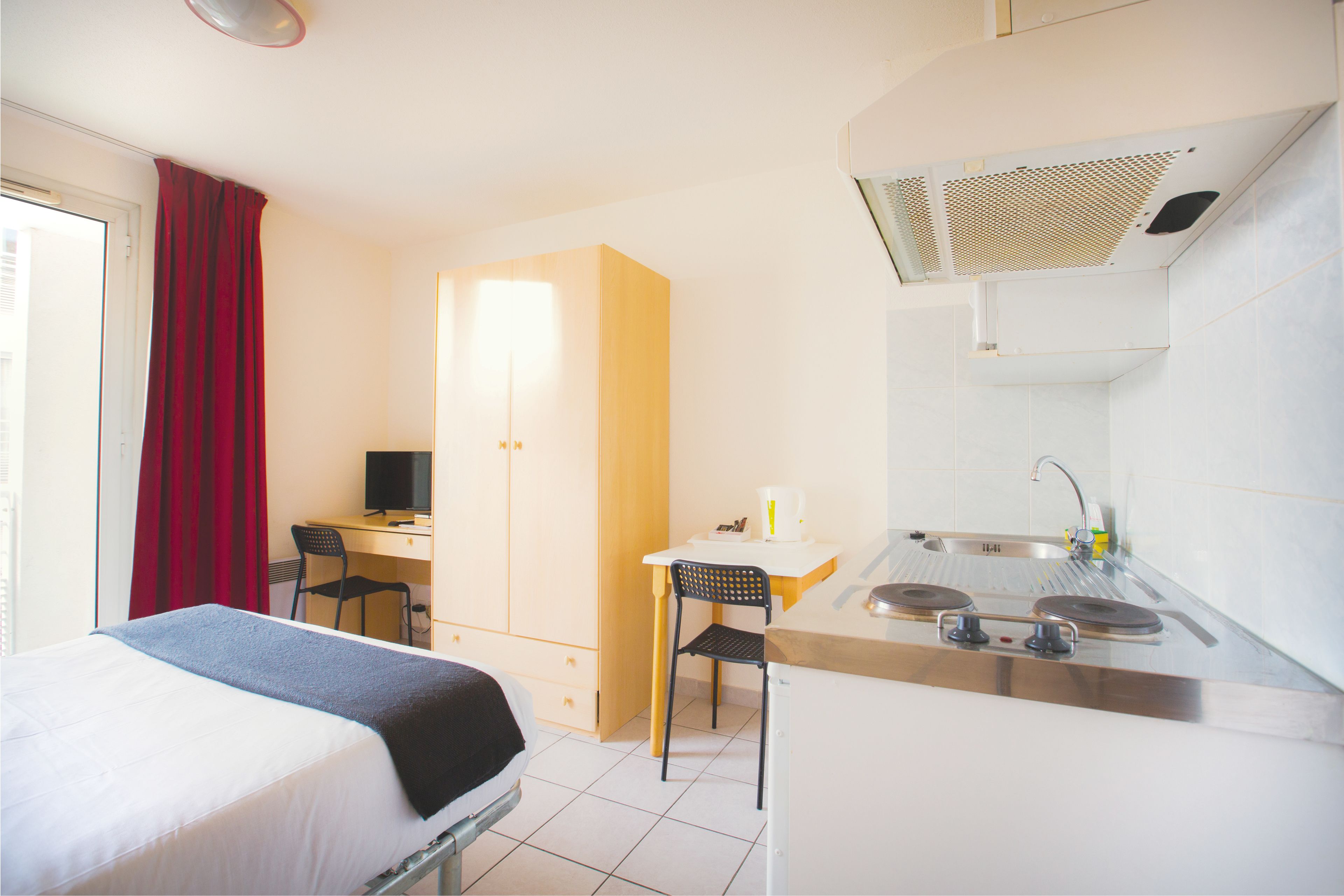 A compact hotel room with a bed, a desk, a chair, a kitchenette, and a window with curtains.