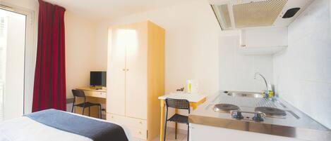 A compact hotel room with a bed, a desk, a chair, a kitchenette, and a window with curtains.